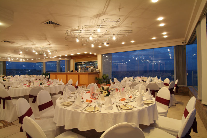 Corporate function room with white chair covers, burgundy sashes and dressed round tables beside panoramic windows